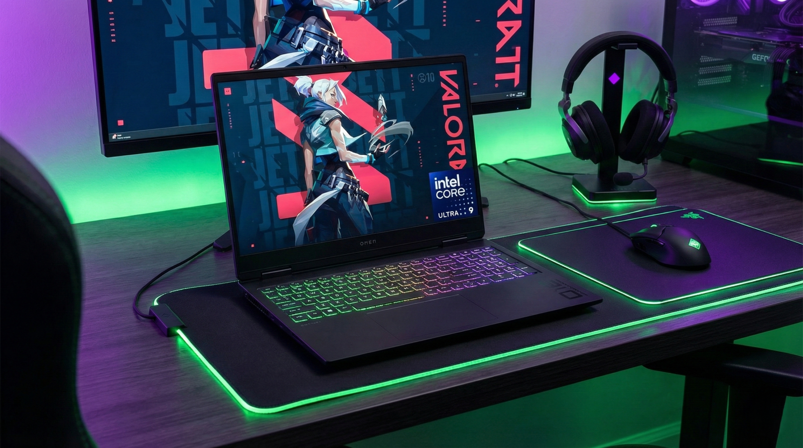 "Portátil gaming HP OMEN MAX 16-ah0010ns abierto en un escritorio de madera. La pantalla muestra el juego Valorant. El setup incluye iluminación LED RGB verde y morada, un monitor externo, auriculares OMEN en soporte y ratón OMEN sobre alfombrilla grande."