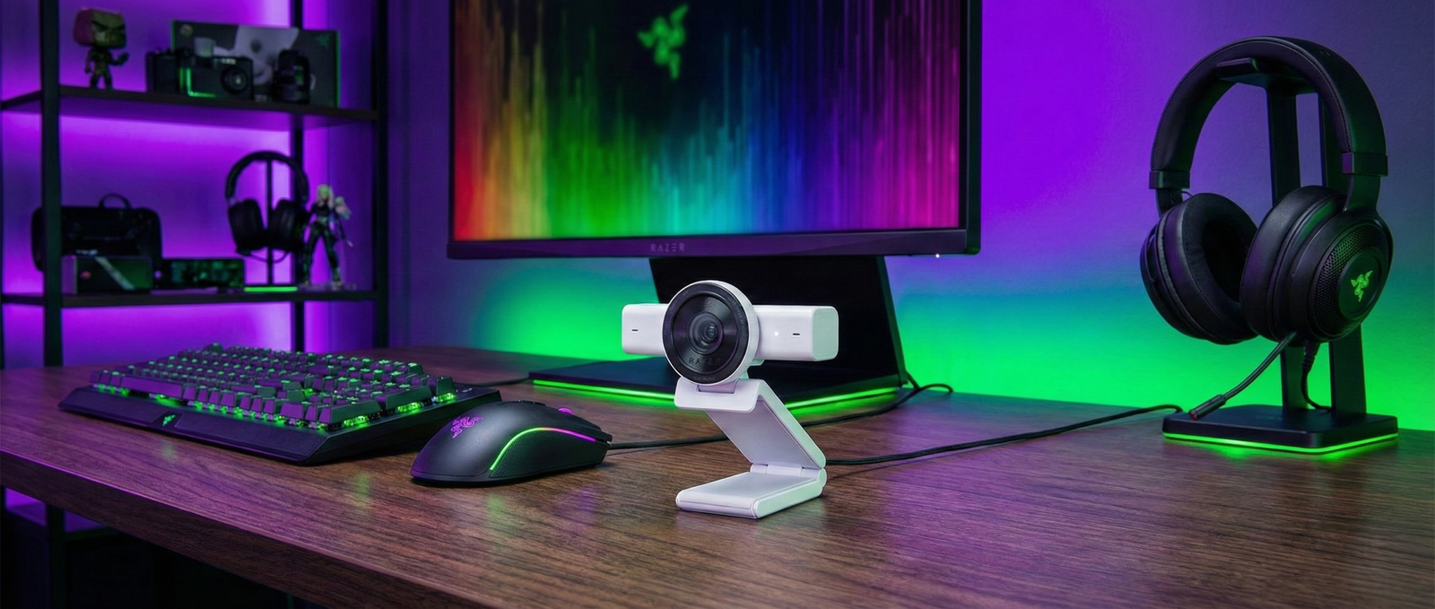 Vista panorámica de un escritorio de streaming completo con la webcam Razer Kiyo V2 4K blanca en el centro, monitor, periféricos gaming y e iluminación ambiental LED en tonos verdes y morados.